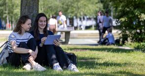 Zwei Studentinnen sitzen im Gras unter einem Baum und schauen auf ein Tablet.