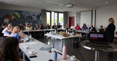 Ein Raum ist mit Menschen gefüllt, die an Tischen in U-Form sitzen. Rechts im Bild hält eine Frau hält einen Vortrag.