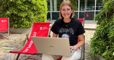 Eine junge Frau sitzt mit ihrem Laptop auf einem roten Liegestuhl und lächelt in die Kamera
