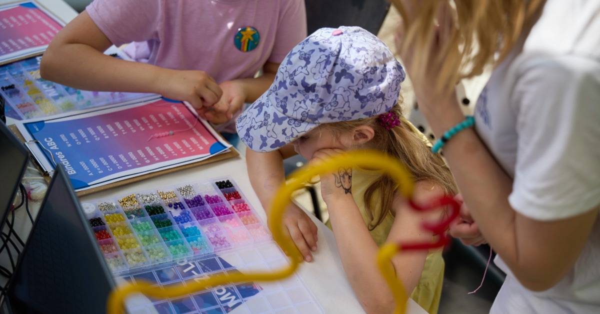 th_luebeck_juniorcampus_perlenketten Kinder sitzen an einem Tisch und fädeln bunte Perlen auf Schnüre. Neben ihnen liegt ein Arbeitsblatt mit einem binären Alphabet.