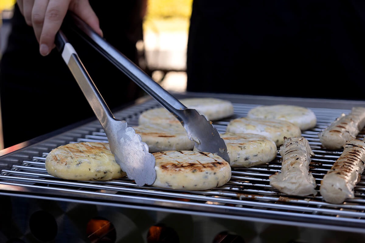 Hand mit Grillzange wendet vegetarische Grillfrikadellen und Bratwürste auf einem Grillrost der Fachschaft Bau