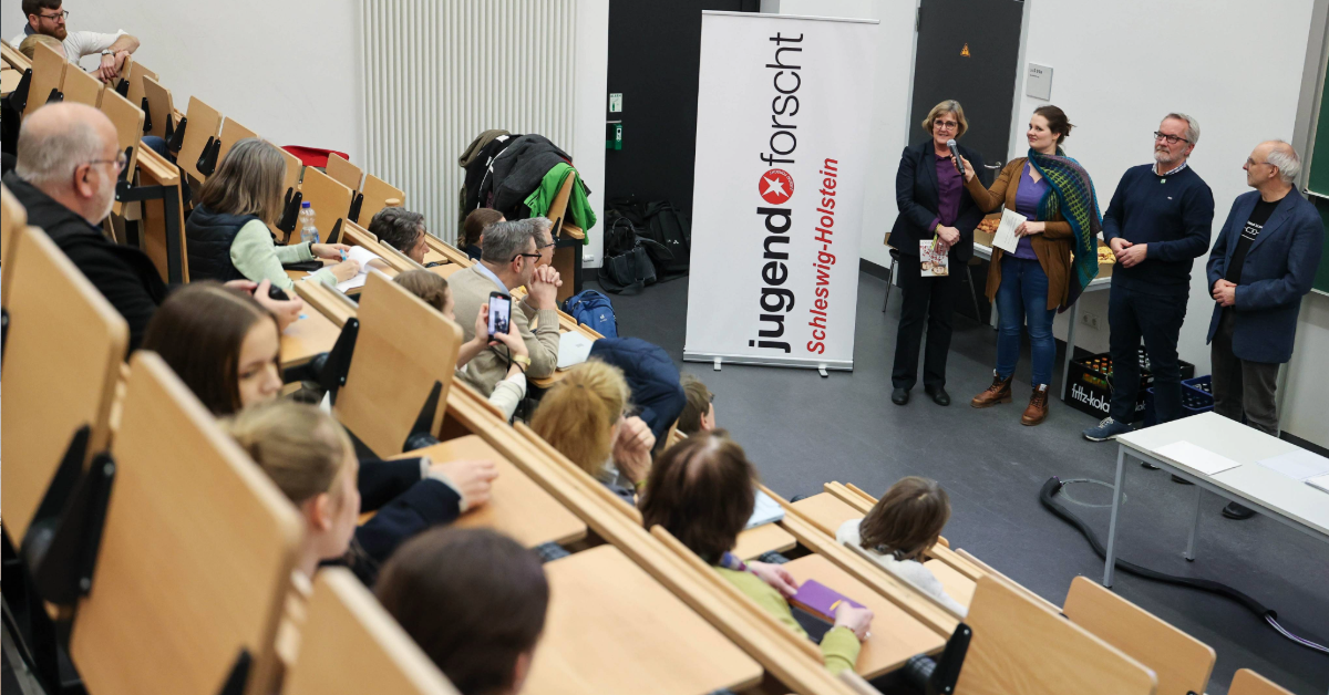Hier sieht man einen Hörsaal und vier Personen, die vorne vor der Tafel stehen