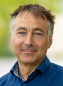Portrait: Aufnahme eines Mannes mit kurzen, dunkelblonden Haaren. Er lächelt und trägt ein dunkelblaues Hemd. Im Hintergrund ist unscharf ein grüner Baum zu sehen.