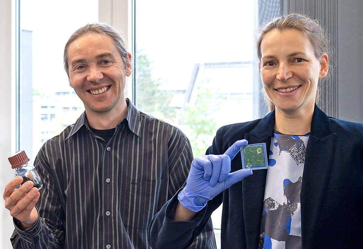 A smiling male and female scientist present their research. The man holds up a small glass vial, while the woman, wearing a blue glove, holds up a small green computer chip.