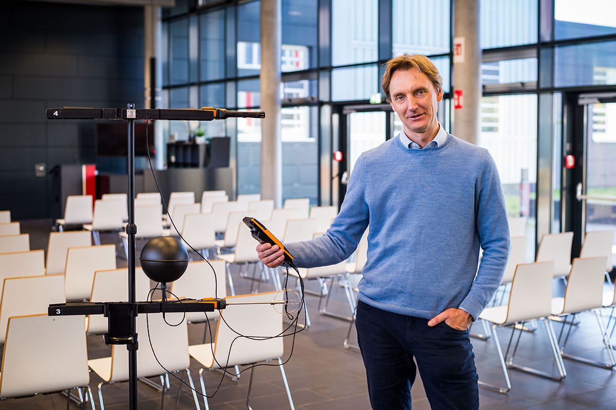 A man in a blue sweater stands in a room with many chairs, holding a technical measuring device.