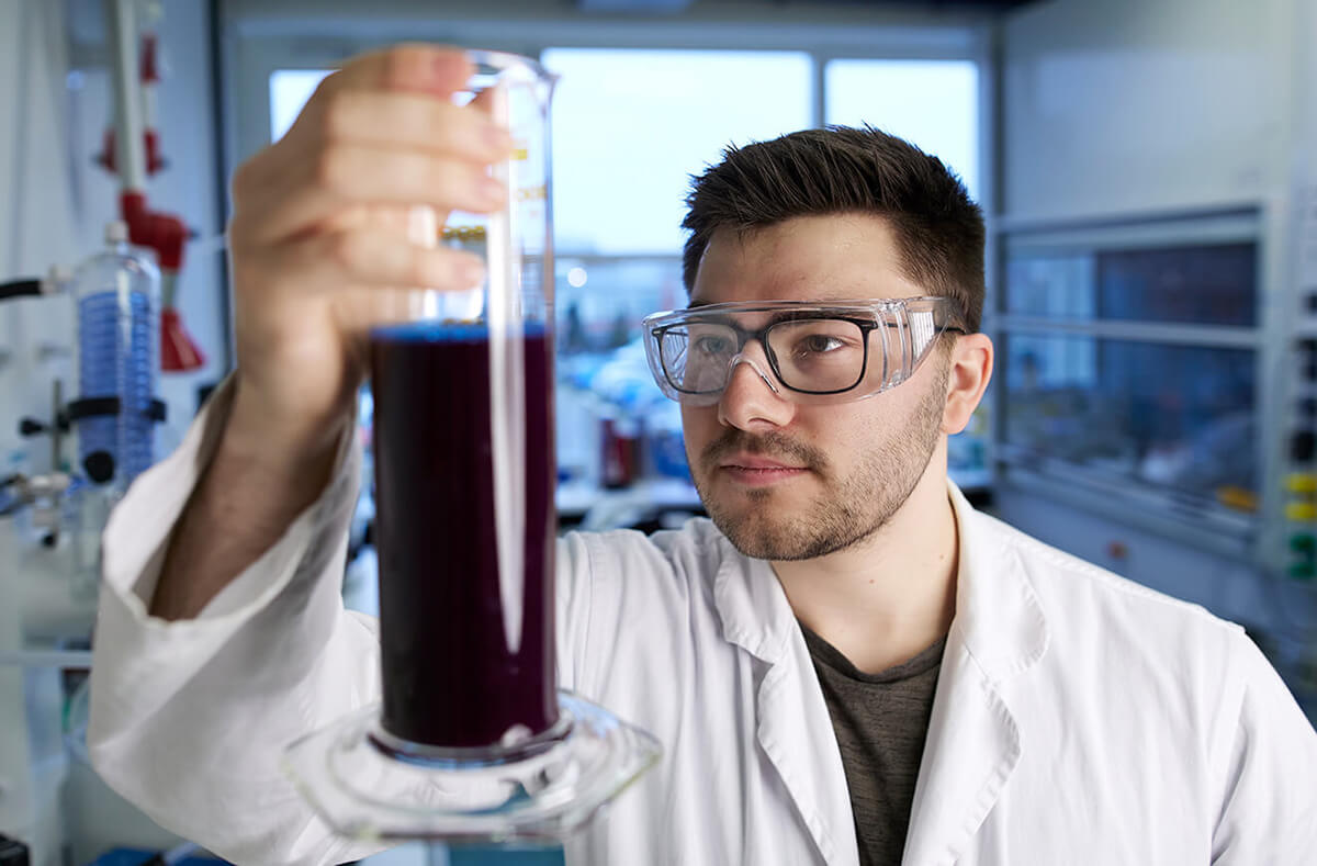 A young male scientist in a white lab coat and safety goggles holds up a graduated cylinder with a dark purple liquid, examining it closely in a laboratory.
