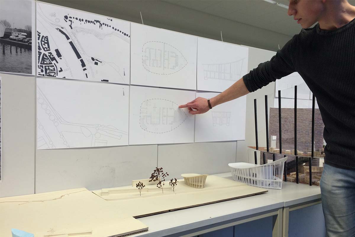 Ein Student zeigt auf einen Entwurfsplan neben Architekturmodellen an einer Wand.