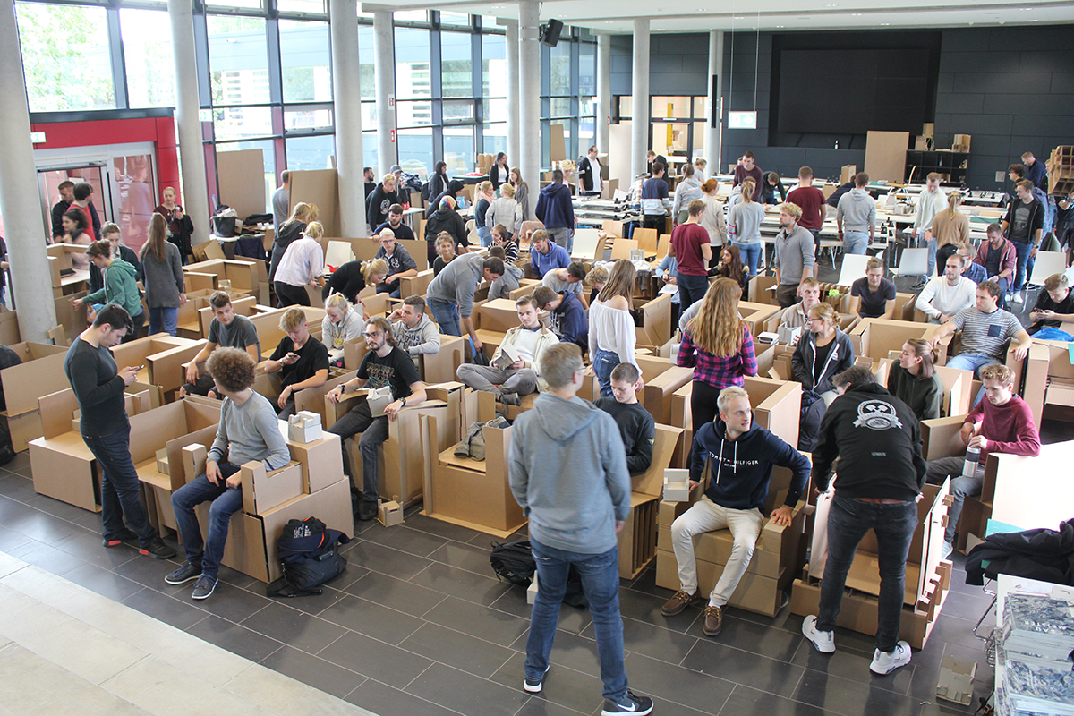 Viele Studierende sitzen auf selbstgebauten Pappstühlen in einer großen Halle.