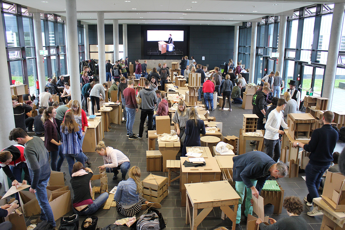 Studierende bauen in einer Halle komplexe Objekte und Möbel aus Wellpappe.