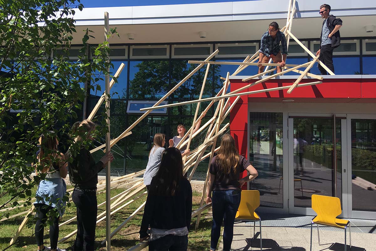 Studierende errichten eine Skulptur aus Holzlatten vor einem Unigebäude.