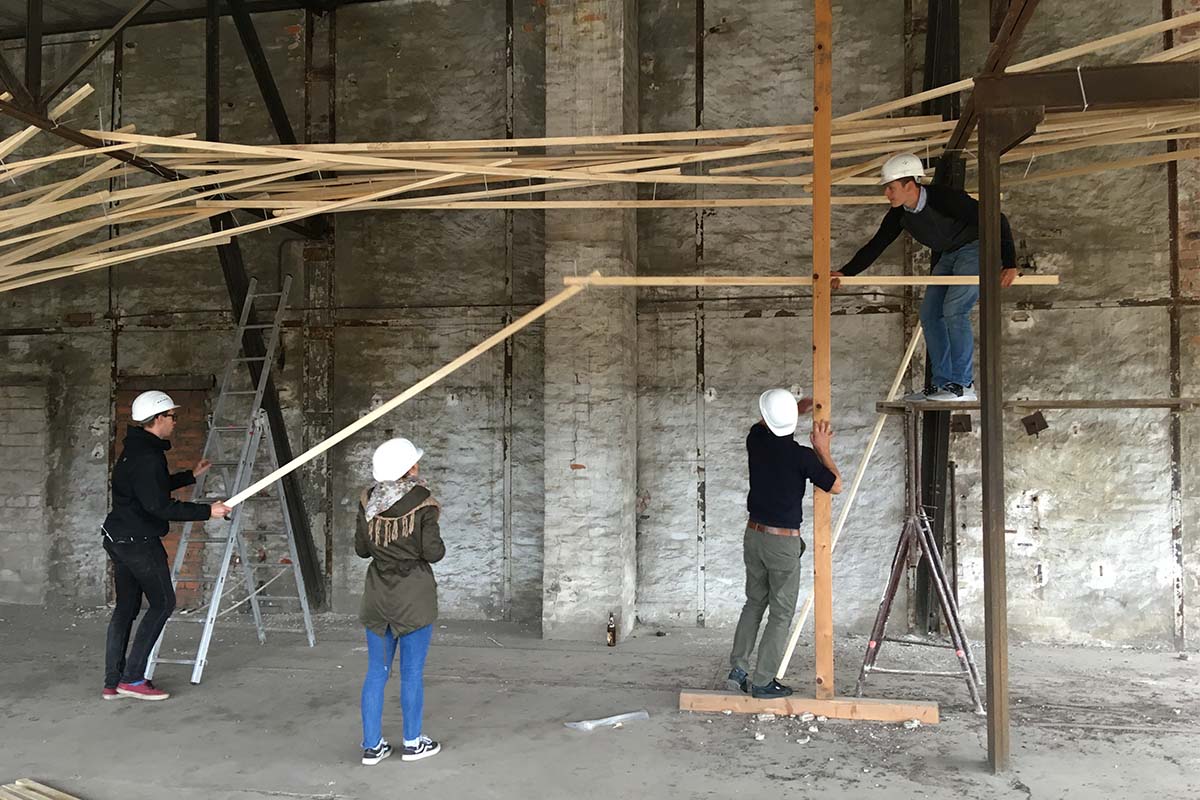 Studierende mit Helmen arbeiten an einer hohen Holzkonstruktion in der Halle.