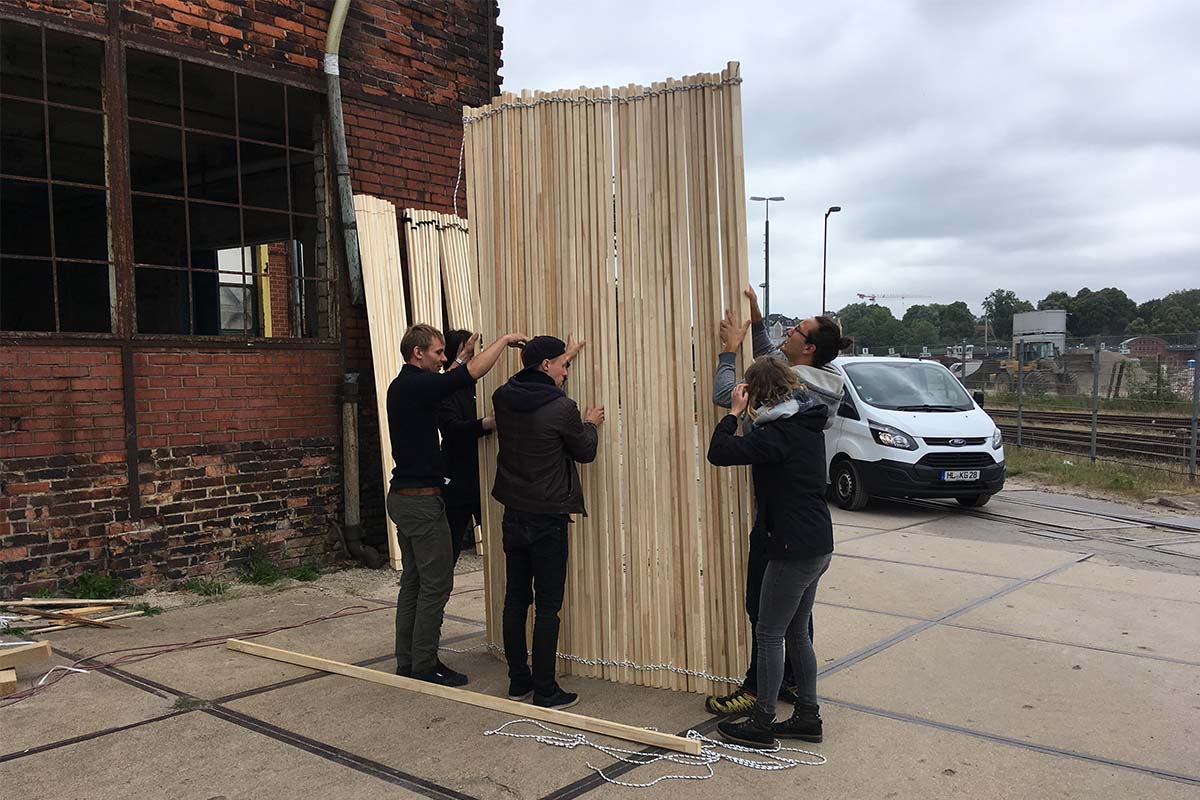 Gruppe trägt ein hohes, geschwungenes Bauteil aus Holzlatten über den Hof.