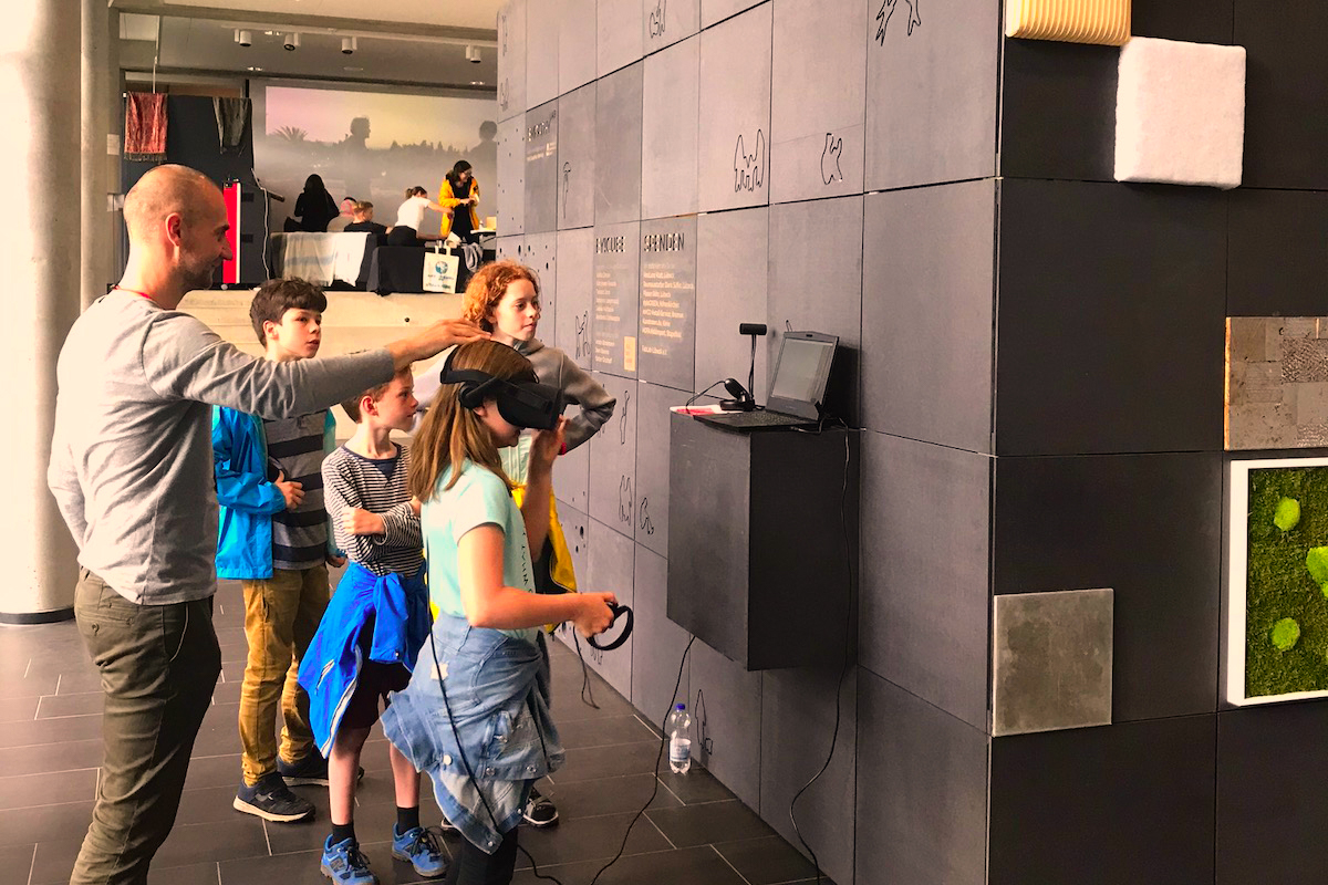 Mann erklärt Kindern VR-Technik, ein Kind nutzt die Brille vor einer Wand.