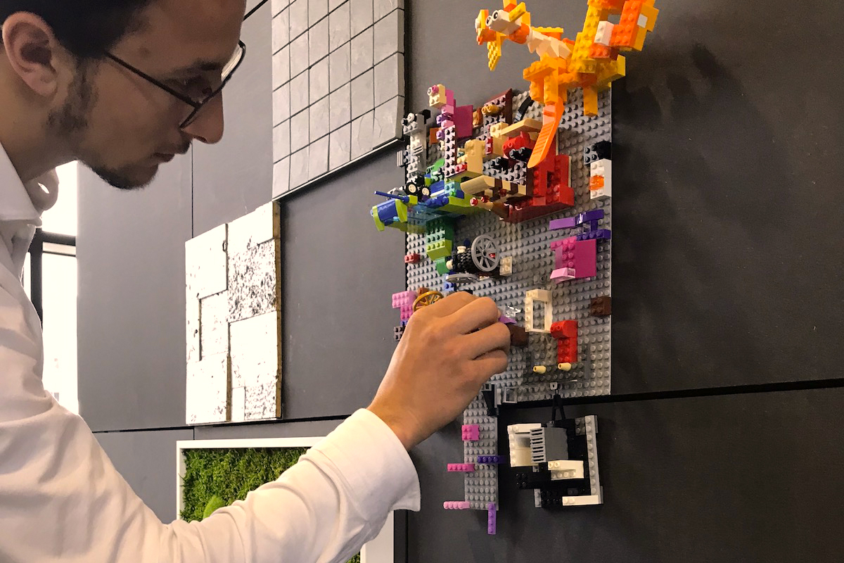 Mann steckt bunte Legosteine an eine vertikale graue Bauplatte.