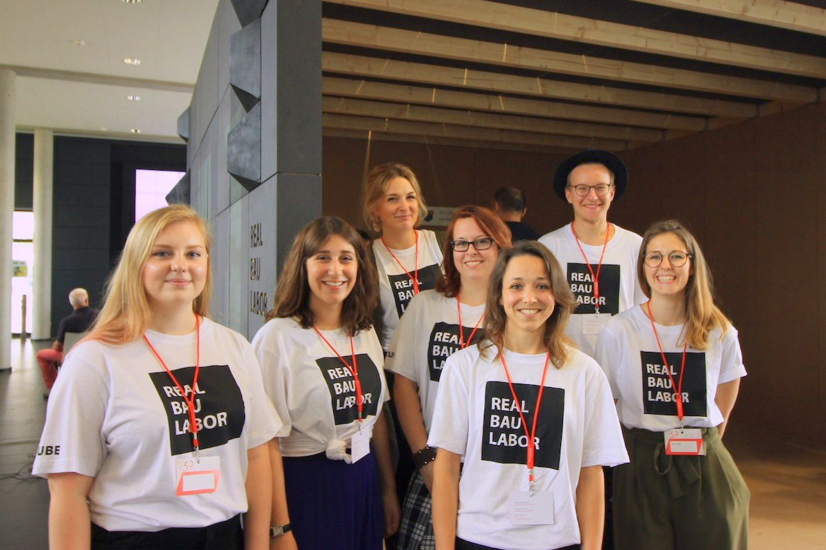 Teamfoto: Sieben Personen in weißen "Real Bau Labor" T-Shirts lächeln.