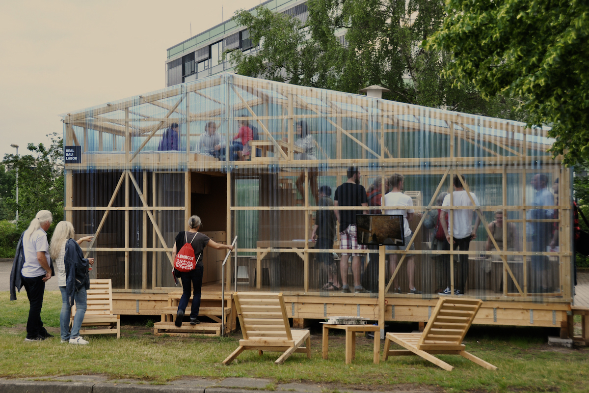 Transparenter Holzpavillon "Real Bau Labor" mit Besuchern auf zwei Ebenen.