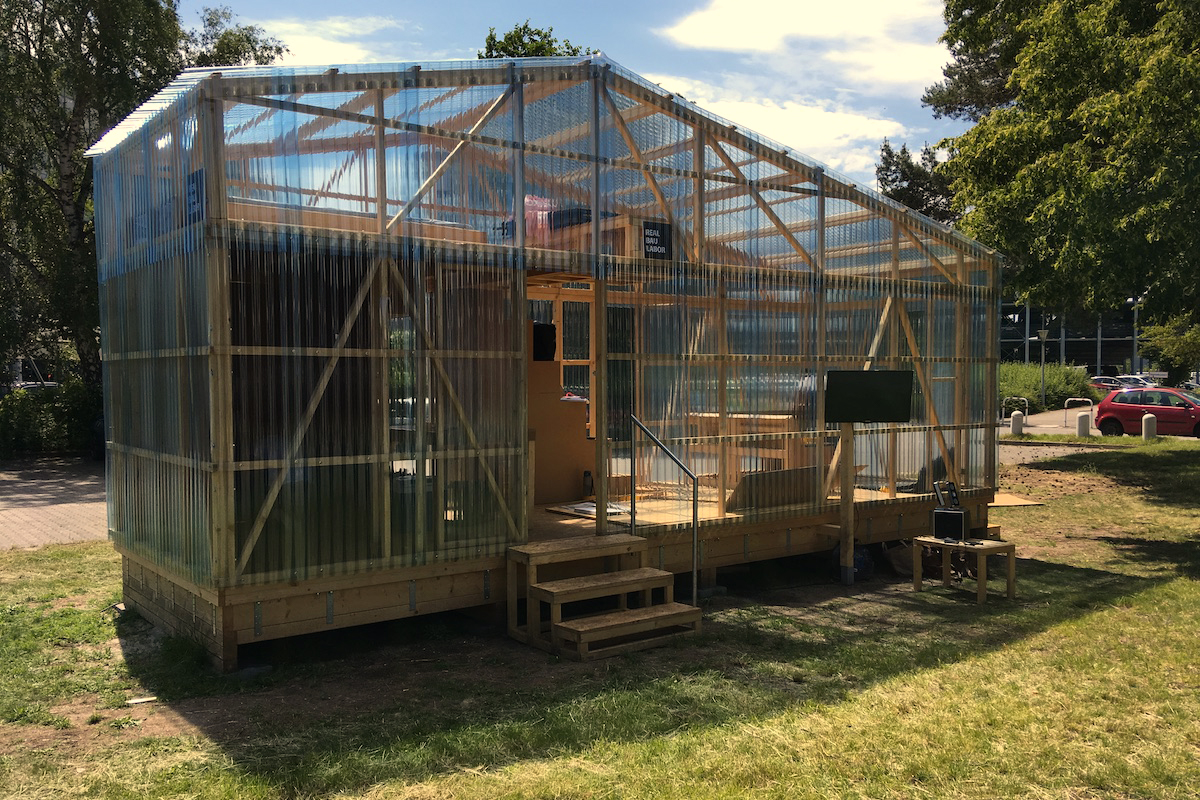 Seitenansicht des transparenten Holzpavillons "Real Bau Labor" im Sonnenlicht.