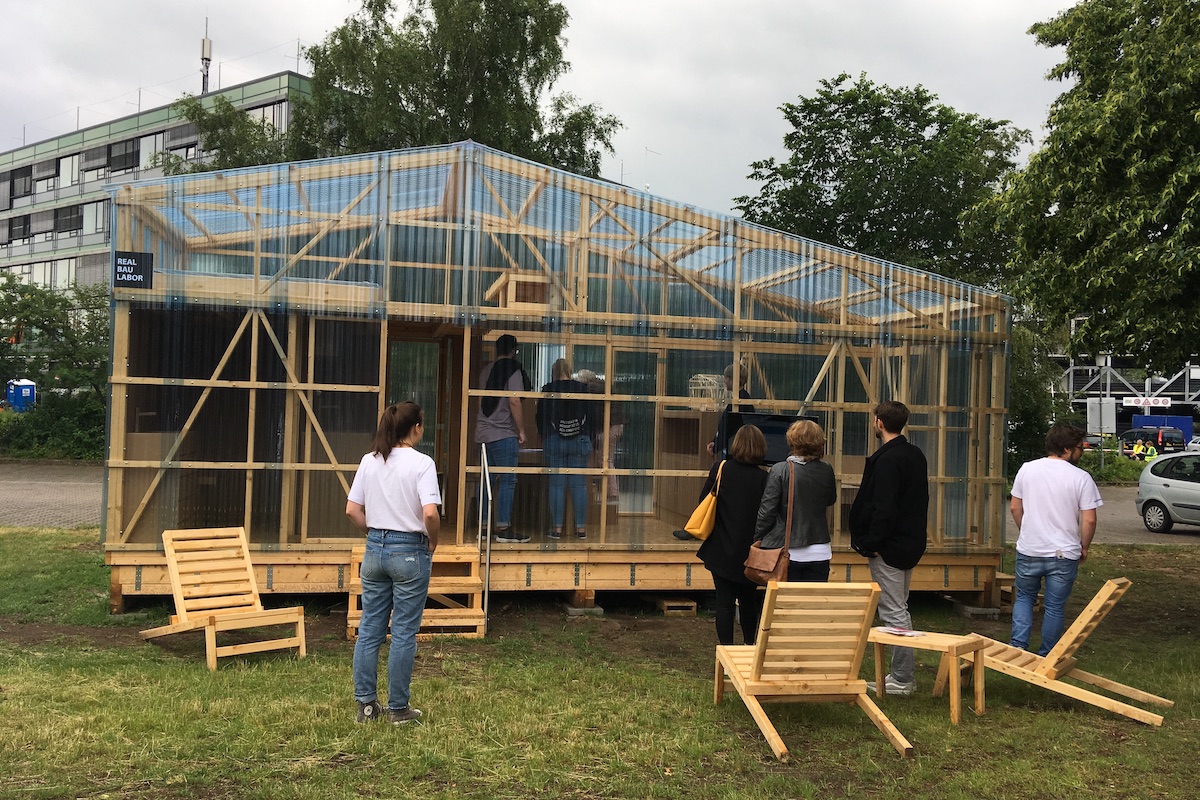 Menschengruppe besichtigt den Holzpavillon "Real Bau Labor" von außen.