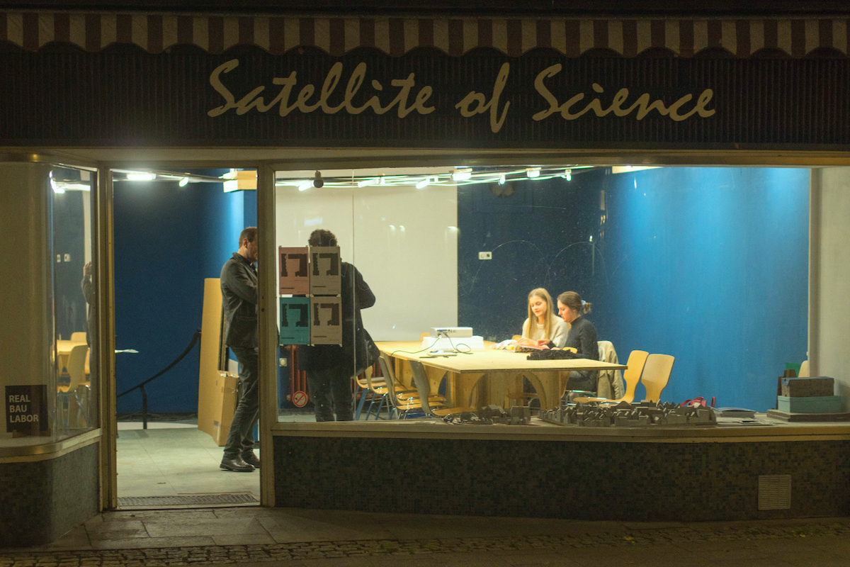 Beleuchtetes Schaufenster "Satellite of Science" nachts, Personen im Innenraum.