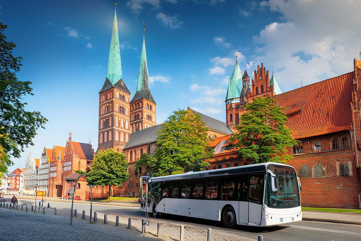 Anreise mit dem Bus Ein weißer Bus fährt auf einer Straße. Im Hintergrund ist die Lübecker Altstadt mit ihren Kirchtürmen und Fachwerkhäusern zu sehen. Grüne Bäume wachsen an der Straße.