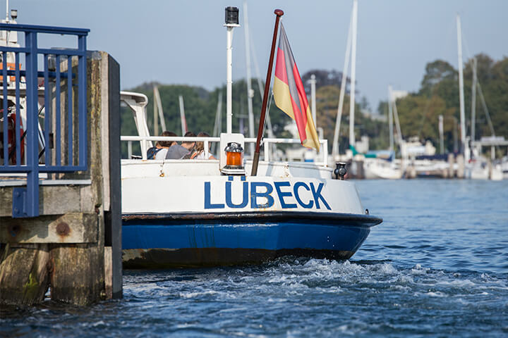Maritimer Blick auf den Travemünder Hafen. Zu sehen ist ein Ausflugsboot mit Personen. Es trägt die Aufschrift Lübeck am Heck.