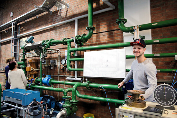 Student mit Cap steht lächelnd vor einem Laboraufbau mit grünen Rohrleitungen und Messgeräten