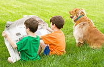 Zwei Kinder sitzen auf einer Wiese neben einem Hund und lesen zusammen in einer Zeitung.