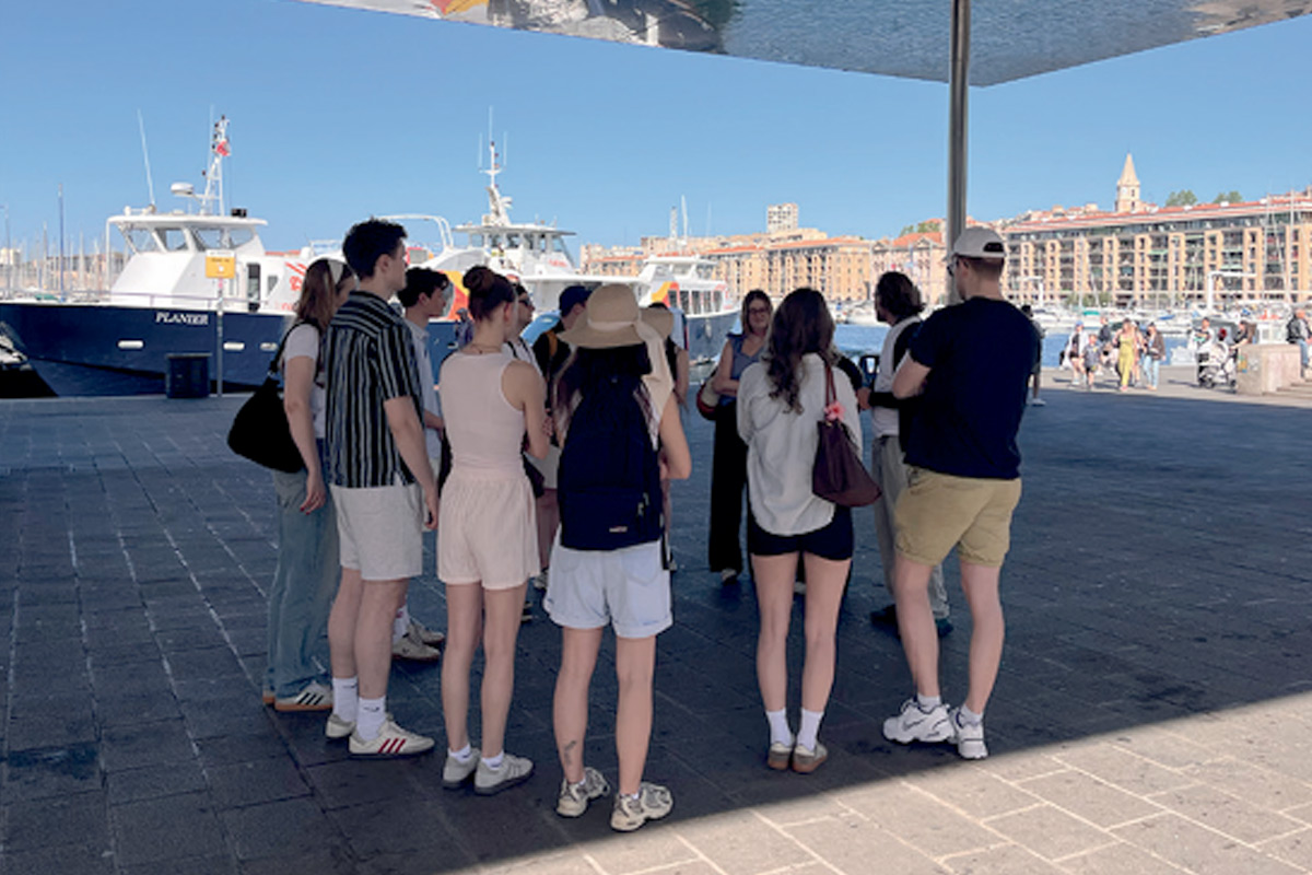 Gruppe von Studierenden der TH Lübeck während einer Exkursion am Hafen von Marseille mit Booten und historischer Stadtkulisse im Hintergrund